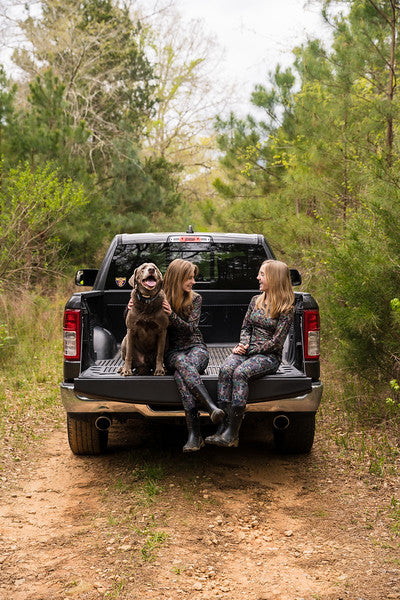 Girl wearing camo hunting clothes sitting on truck tailgate with dog.