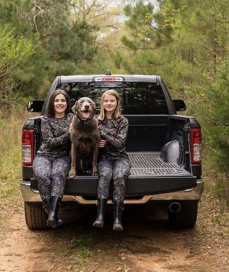 girls sitting on truck tailgate with dog. Girls wearing hunting clothes.